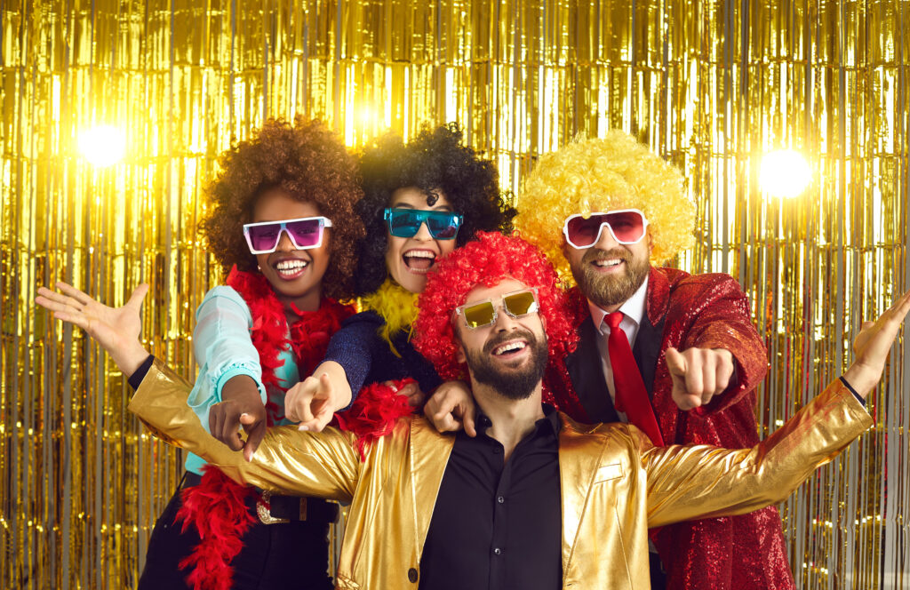 Group of friends posing in colorful wigs and 70s disco costumes at a party.
